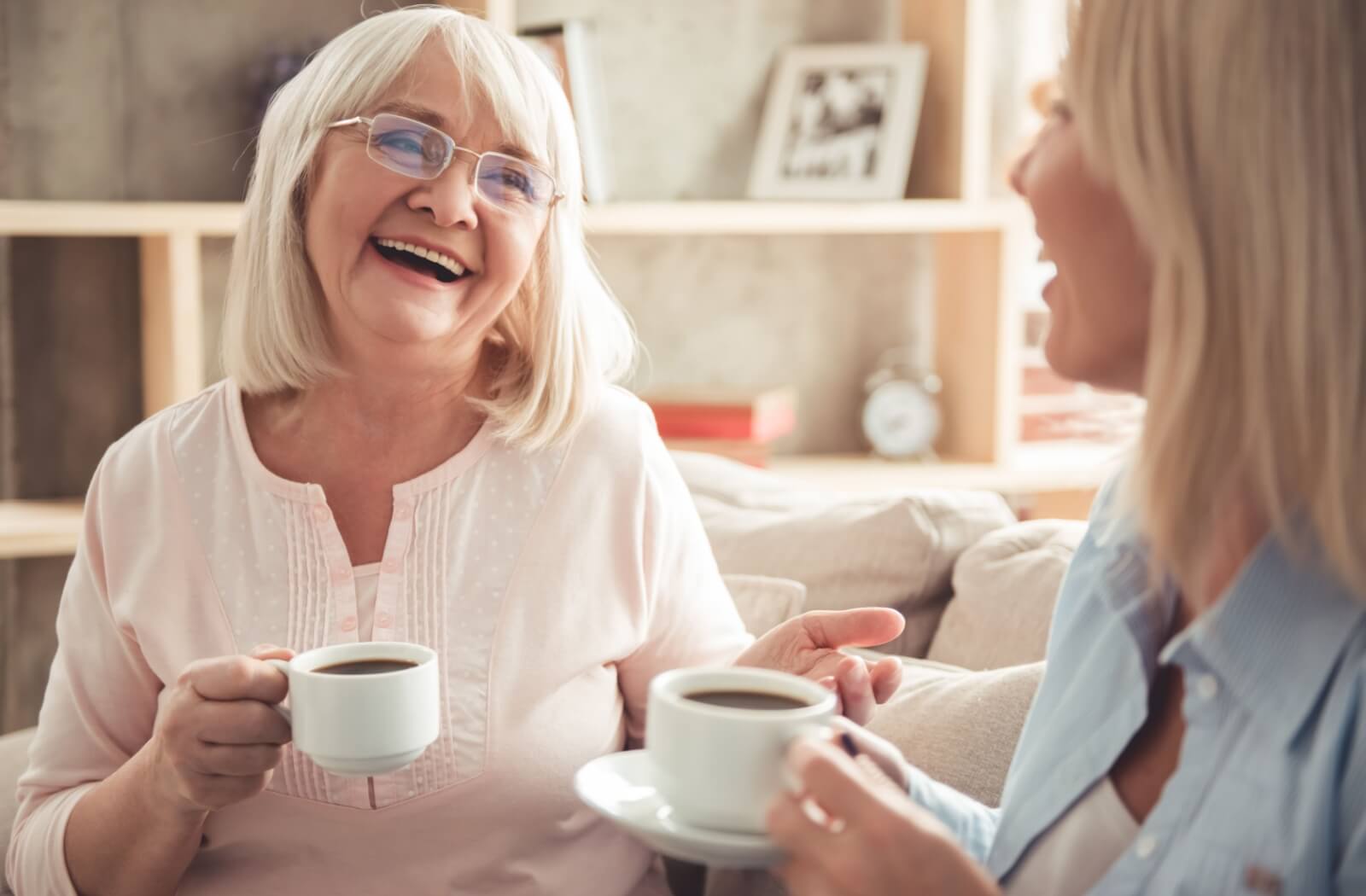 A senior parent and their older adult child sitting on a couch smiling and talking to each other while each holding a cup of tea