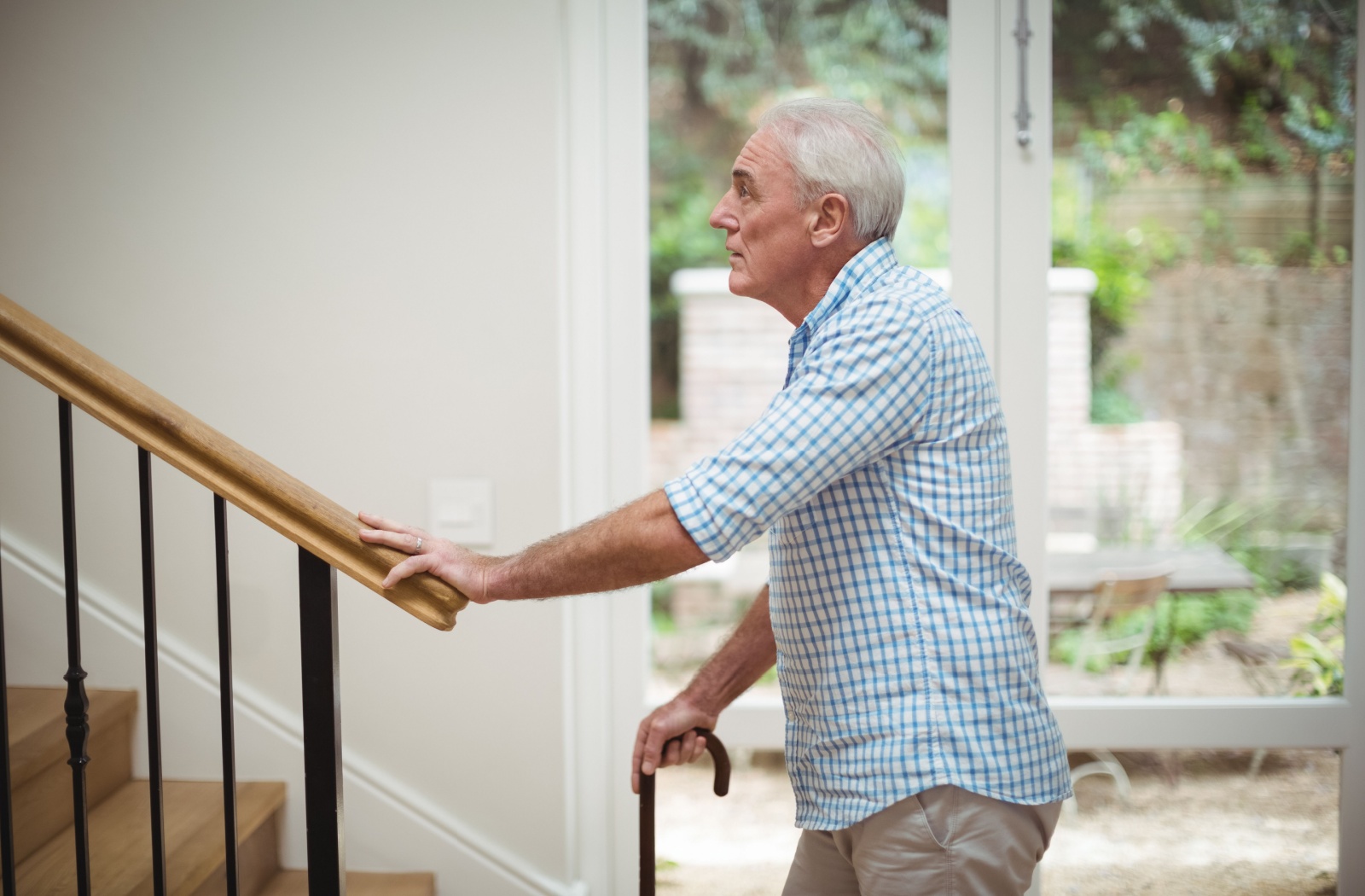 An older adult with a walking cane in his right hand stands at the bottom of a staircase looking up with his left hand on the banister.