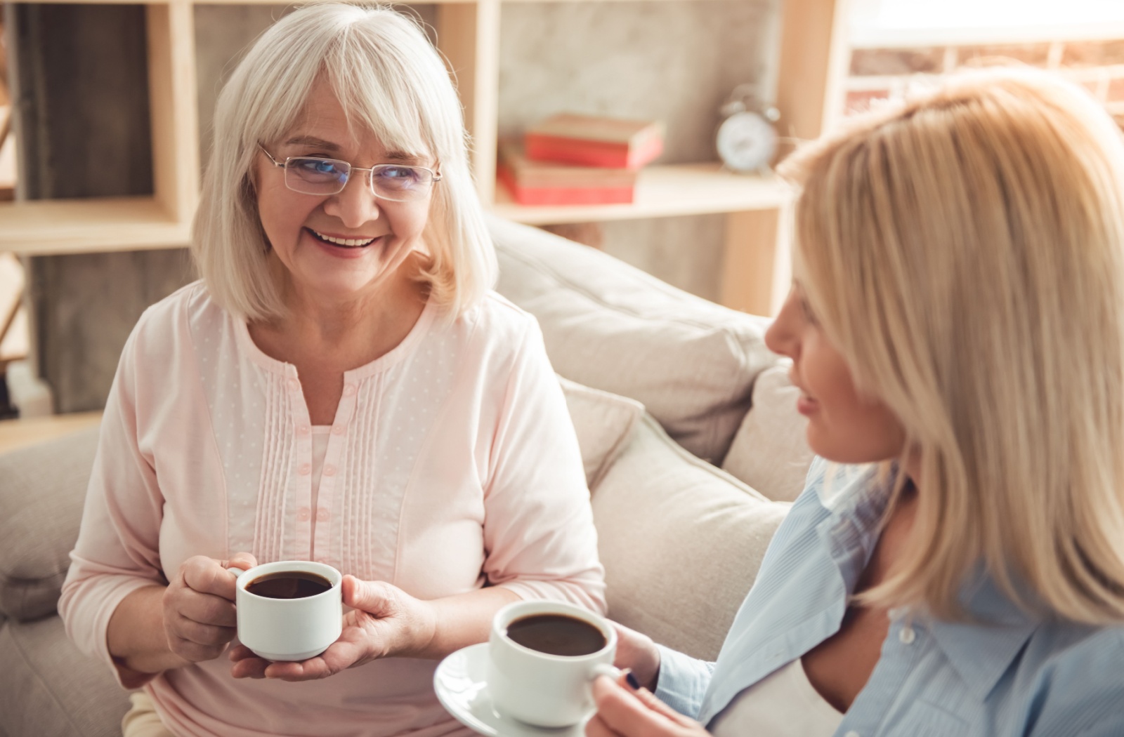 A senior mother and her adult daughter sitting on a couch drinking coffee and smiling
