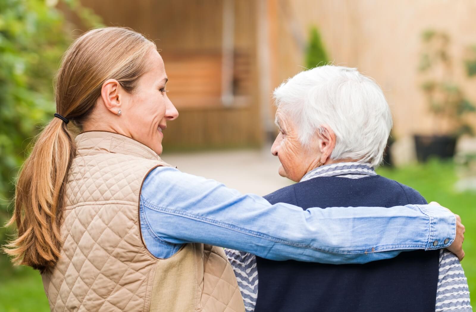 An adult child hugging their parent with dementia from the side and smiling as they walk together outside.