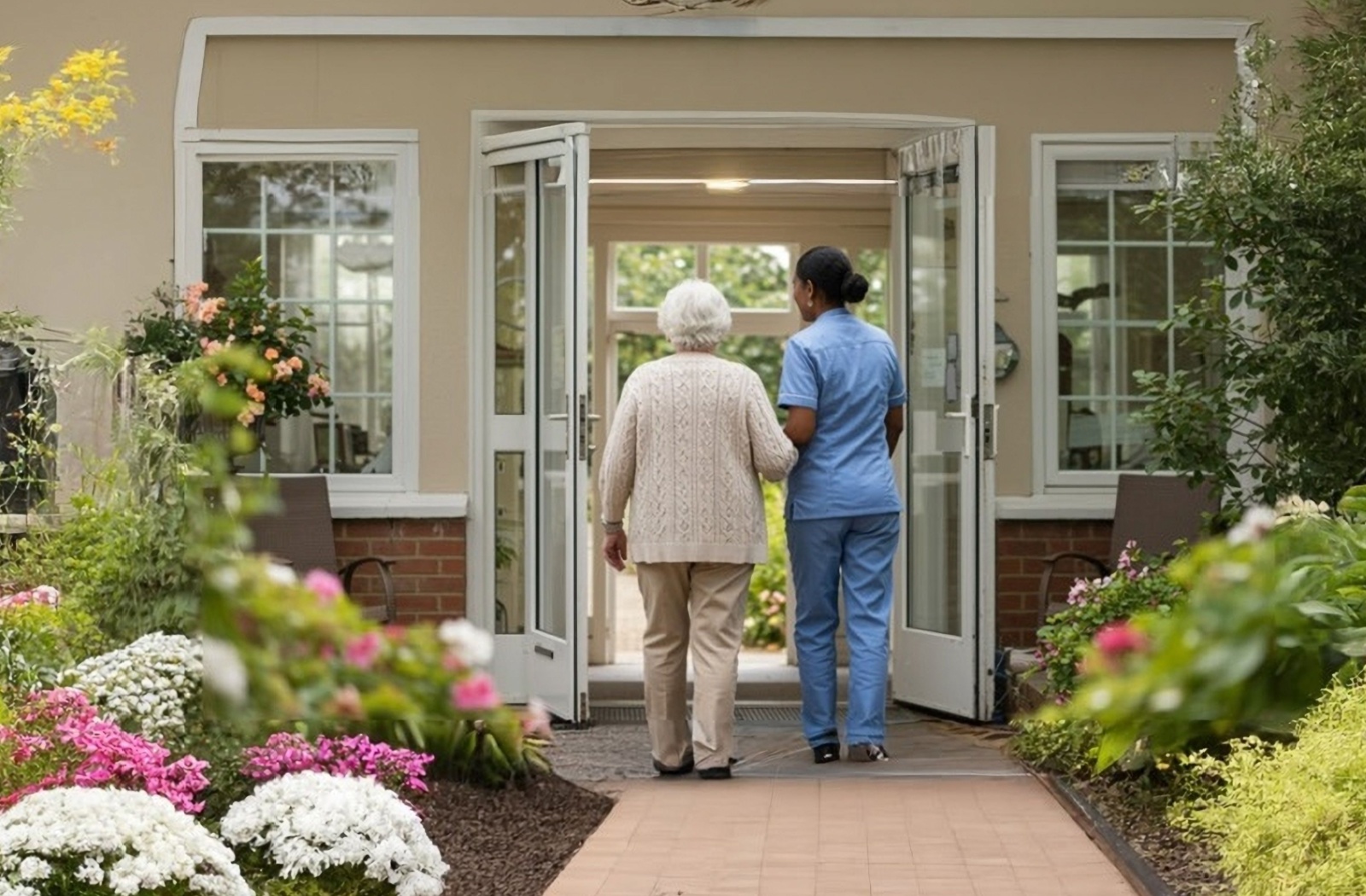 Senior walking with caregiver toward memory care community entrance surrounded by flowers and greenery