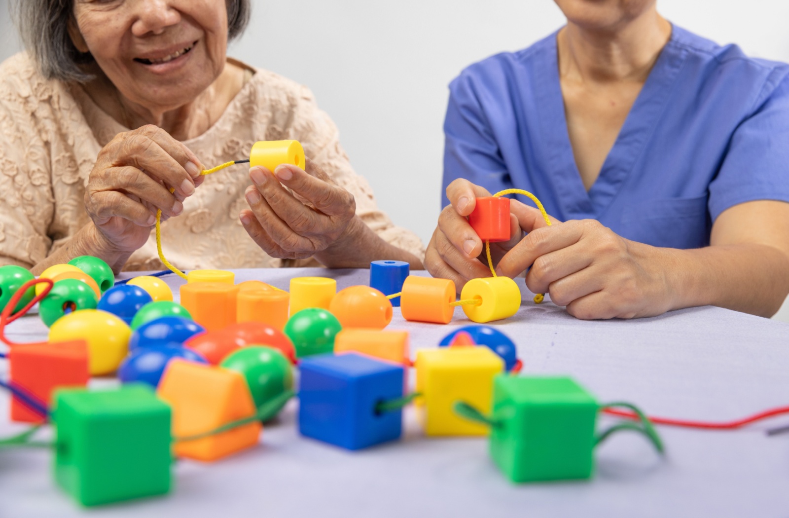 A nurse sits with a senior while completing a puzzle of putting blocks on a string to help support cognition in memory care