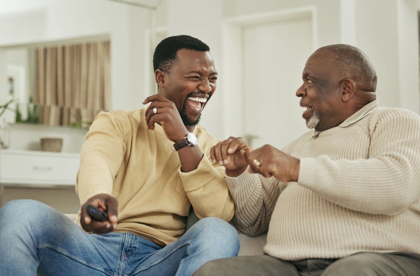 A senior and their adult child laugh together while watching TV on a couch