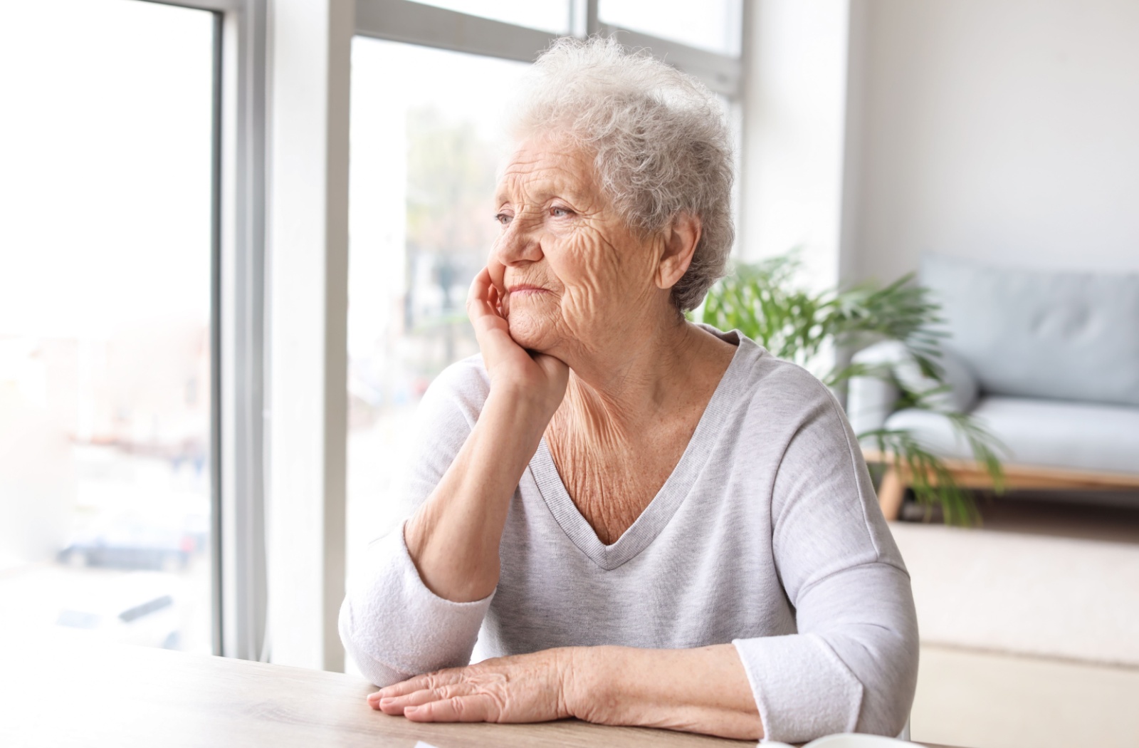 A senior living with dementia gazes out a window in a bright living room.