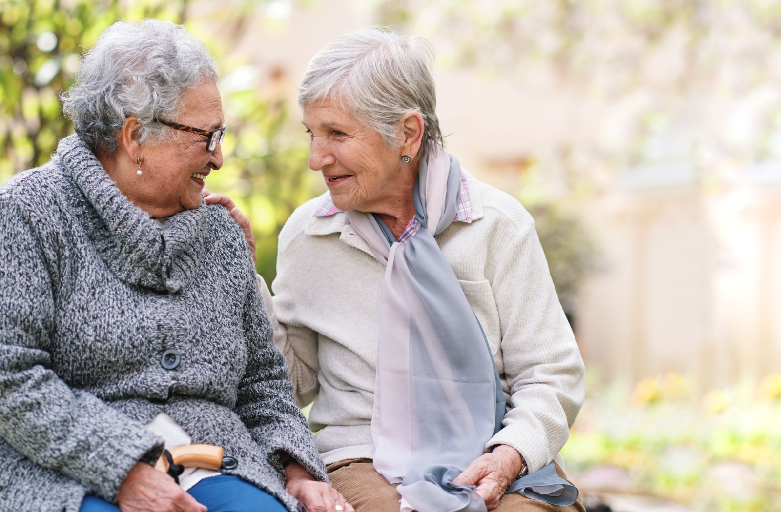 Two older adults smile and converse while sitting on a bench in the garden of their senior living community