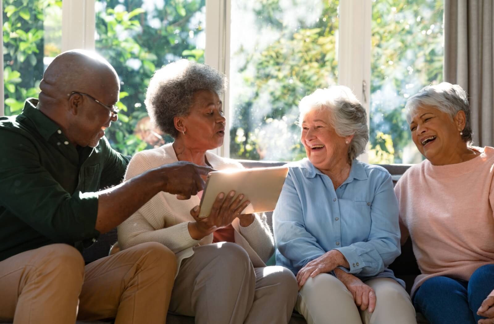 A group of 4 older adults laugh while flipping through a photo together underneath a window in personal care

